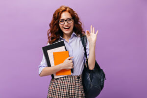 Red haired lady in eyeglasses holds books and shows ok sign. Ginger school girl with curly hair in lilac shirt and plaid skirt smiling..